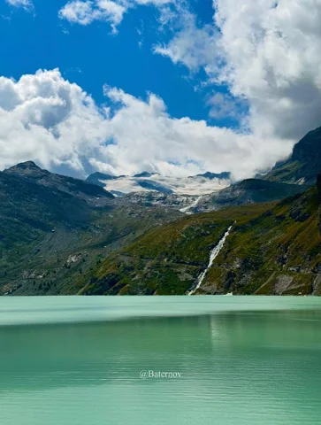 Heller Bergsee neben einem Wasserfall und wolkenverhangenen Gipfeln