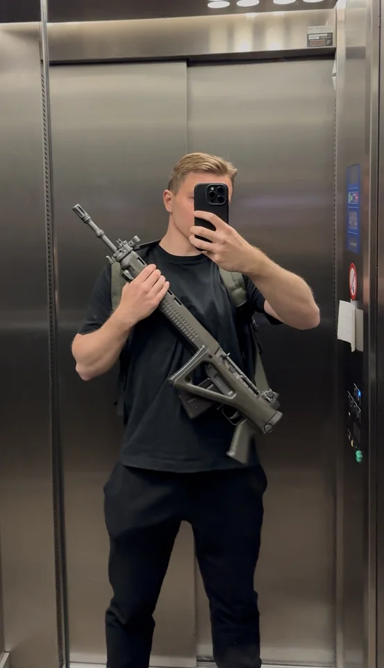 Brandon haciéndose una foto en el espejo de un ascensor mientras sostiene un fusil.